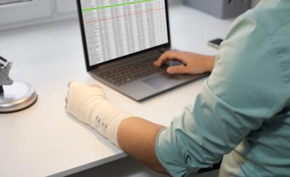 Mężczyzna z kontuzją ręki pracujący na laptopie w biurze Mężczyzna z bandażem owiniętym wokół uszkodzonego nadgarstka, siedzący przy biurku i korzystający z arkuszy biznesowych na nowoczesnym notebooku