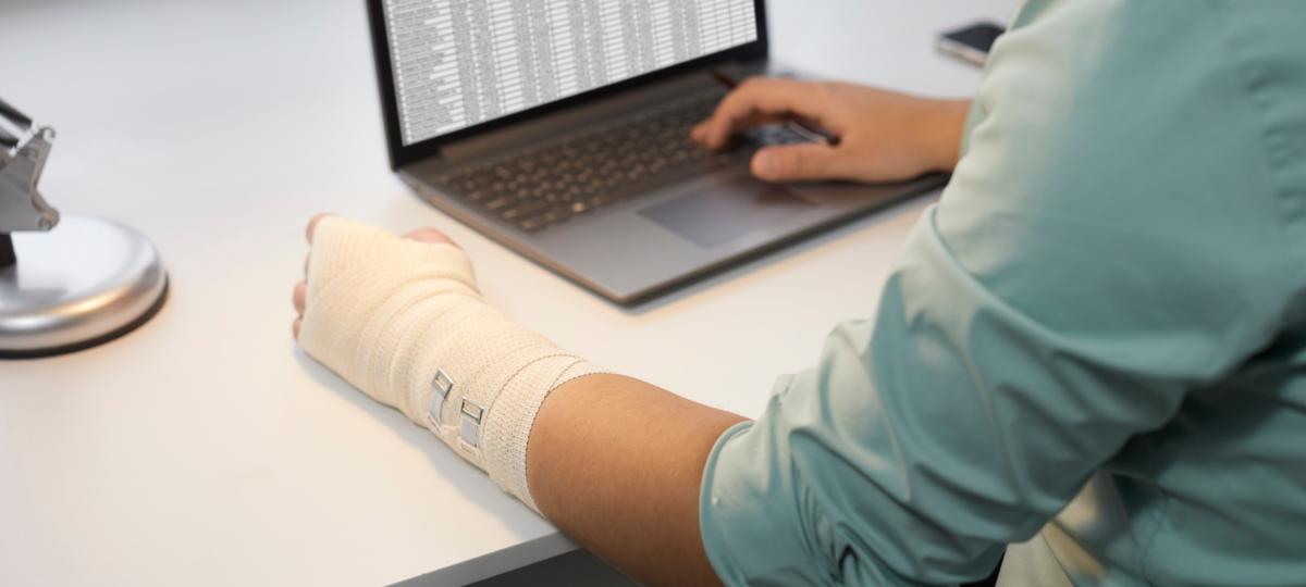 Mężczyzna z kontuzją ręki pracujący na laptopie w biurze Mężczyzna z bandażem owiniętym wokół uszkodzonego nadgarstka, siedzący przy biurku i korzystający z arkuszy biznesowych na nowoczesnym notebooku